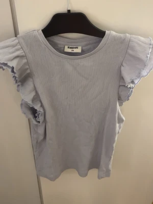 Ljuslila ribbad topp med volangärm - Säljer en ljuslila ribbad topp från Kappahl i storlek 170. Toppen har rund hals och ärmlös design med breda volangärmar som ger en cool och feminin touch. Perfekt att styla med jeans eller kjol för en fräsch look.
