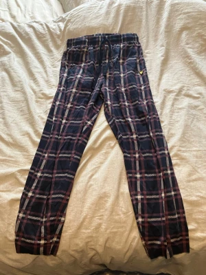 Rutig mjukisbyxa från Lyle & Scott - Säljer ett par mjukisbyxor från Lyle & Scott i storlek S. Byxorna har ett mörkblått och vinrött rutigt mönster, snörning i midjan och ett broderat gult logomärke på sidan. Materialet är mjukt bomullstyg som är skönt att ha på sig.