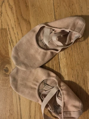 Rosa balettskor med resårband - Säljer ett par klassiska balettskor i ljusrosa bomull med elastiska band över foten för extra stöd. Skorna har rund tå och mockadetaljer på sulan för bättre grepp. Perfekta för danslektioner eller träning. Har köpt begagnat och har används flera gånger.