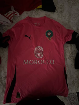 Marocko fotbollströja Puma rosa - Säljer en rosa fotbollströja från Puma med Marockos landslagsemblem på bröstet och trycket 'MOROCCO' i silver. Tröjan har korta ärmar och rund hals, tillverkad i lätt polyester som andas. Perfekt för dig som gillar fotboll och vill sticka ut med färg.