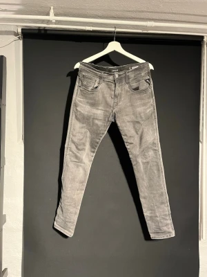 Replay grå jeans slim fit, storlek 29 - Snygga grå jeans från Replay i slim fit-modell, storlek 29. Jeansen har klassisk femficksdesign, svarta detaljer och Replay-logga på bakfickan. Materialet är mjukt och stretchigt jeans-tyg som sitter skönt. Perfekt för dig som gillar en modern och stilren look. Jeansen har en skavank vid fickan men inget man tänker på vid användning (därav billigt pris)