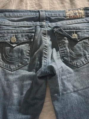 True Religion Bootcut Jeans ljusblå - Säljer ett par ikoniska ljusblå True Religion jeans med bootcut passform. Jeansen har snygga kontrastsömmar, stora fickor med lock och silverfärgade knappar. Klassisk denimlook med tydliga detaljer och broderad logga på bakfickorna. De är i storlek 27(Xs/S) Säljer för att de är för stora för mig. Passar någon som är 160 då är de lite korta. Köpte på vinted och har bara provat hemma!