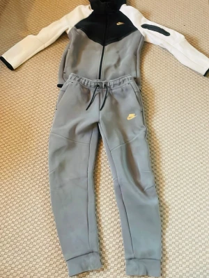 Grå, svart och vitt Nike tech - Bilderna visar ett Nike-set med hoodie och joggers i grått, svart och vitt, men inga skor finns med. Setet har guldfärgade Nike-loggor och är gjort i syntetmaterial med dragkedja och snörning på byxorna. Storleken är 147-158 cm och har ett litet håll bak på byxorna.
