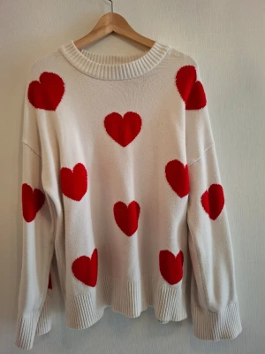 Vit stickad tröja med röda hjärtan FB Sister - Säljer en vit stickad tröja från FB Sister i storlek M. Tröjan har ett gulligt mönster med stora röda hjärtan över hela och breda ribbade muddar vid hals, ärmslut och nederkant. Perfekt för dig som gillar statement-plagg och vill ha något mysigt och snyggt. Förekommer en liten fläck.
