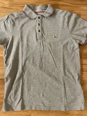 Moncler Pike - En riktigt stilig och klassisk Piké från Moncler i färgen grå. Tröjan är äkta och i bild 2 ser man att den är autentiskt. Perfekt för festligare tillfällen! 