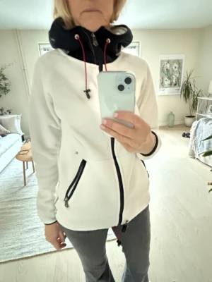 Vit fleecejacka från Pelle P - Säljer en vit fleecejacka från Pelle P i storlek XS. Jackan har svart hög krage med snörning i rött, hel dragkedja och två fickor med svarta dragkedjor. Perfekt för kyliga dagar och har en sportig look med snygga kontrastdetaljer.