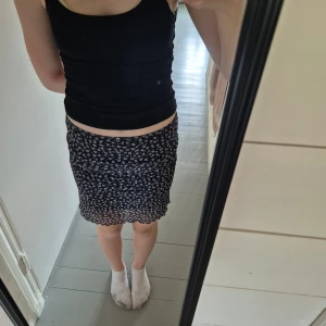 Svart blommig kortkjol från Monki - Säljer en svart kortkjol från Monki med vitt blommigt mönster. Kjolen har en figurnära passform och är tillverkad i ett lätt, stretchigt material. Nederkanten har en vågig volangdetalj som ger en söt touch.🫶