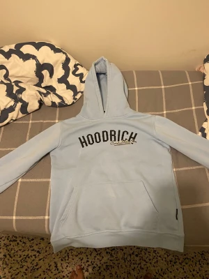 Ljusblå hoodie från Hoodrich - Snygg ljusblå hoodie från Hoodrich med svart och vit logga på bröstet och tryck på ryggen. Tröjan har huva, magficka och långa ärmar. Perfekt för streetwear och chill. Mjuk bomullskänsla och normal passform.