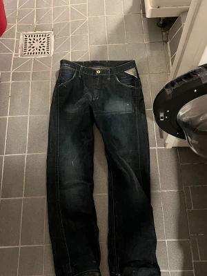 Replay jeans med gul brodyr och dragkedjor - Säljer ett par mörkblå Replay jeans med snygga guldfärgade broderier och dragkedjor på bakfickorna. Klassisk femficksmodell med raka ben och normal passform. Jeansen har gul knapp och detaljer som sticker ut. Perfekt för dig som gillar street och vill ha något unikt.