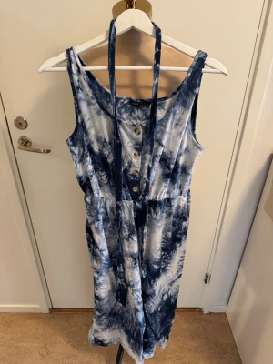 Blåvit batikklänning från SHEIN - Snygg jumpsuit från SHEIN i blåvitt batikmönster. Jumbsuit är ärmlös med knappar framtill och har ett knytband i midjan. Tillverkad i mjuk bomull, perfekt för sommarens varma dagar.