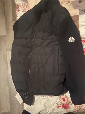 Svart pufferkofta från Moncler - Snygg svart pufferjacka från Moncler med stickade ärmar och klassisk logga på armen. Jackan har quiltad front, dragkedja och ribbade detaljer. Perfekt för dig som vill ha en stilren och varm jacka med exklusiv känsla och modern passform. Notera att dragkedjan är lite seg och kan bli svår att dra upp. Den är S men kan också passa xs