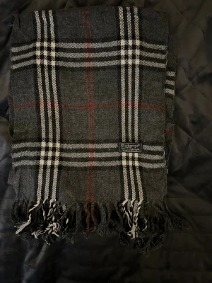 Grå rutig halsduk från Burberrys - Snygg halsduk i klassiskt rutigt mönster från Burberrys. Färgerna är grå, svart, vitt och tunna röda linjer. Gjord i 100% lammull och har fransar i ändarna. Perfekt accessoar för att lyfta din outfit med en tidlös brittisk touch.