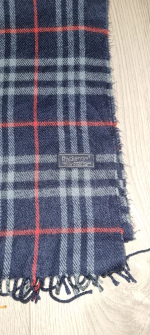 Rutig ullhalsduk från Burberrys - Klassisk rutig halsduk från Burberrys i marinblått, grått och rött. Tillverkad i 100% lammull med fransar i ändarna. Perfekt accessoar för att lyfta din vinterstil med en tidlös brittisk känsla.