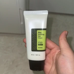 COSRX Aloe Soothing Sun Cream SPF50 - Aloe Soothing Sun Cream från COSRX med SPF 50 PA+++ i en vit tub med svart lock. Innehåller aloe vera-extrakt som återfuktar och skyddar mot UVA och UVB. Tuben rymmer 50 ml och är perfekt att ha med sig i väskan för dagligt solskydd.