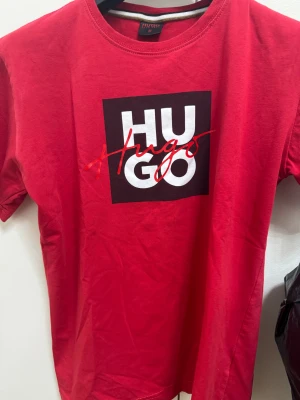 Röd HUGO t-shirt med tryck - Snygg röd t-shirt från HUGO i bomull med klassisk rund hals och korta ärmar. Framsidan har ett stort svart och vitt tryck med HUGO-loggan och röd handskriven text över. Perfekt för dig som gillar streetstyle och vill sticka ut.