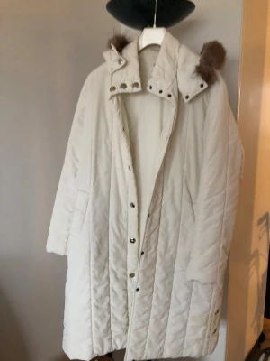 Vit lång pufferjacka med huva - Säljer en vit pufferjacka med huva och snygga bruna fuskpälsdetaljer. Jackan har quiltad design, tryckknappar framtill och två stora fickor. Perfekt för kalla dagar med sin långa och loose passform. Stilsäker och varm, passar dig som gillar enkel men trendig look.