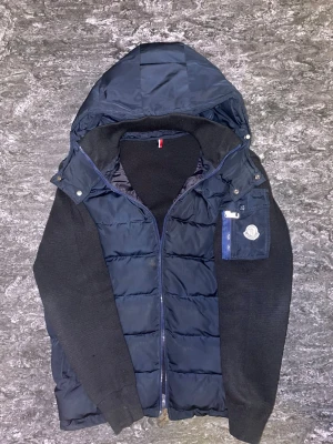 Svart och mörkblå pufferjacka Moncler - Moncler pufferjacka i svart och mörkblå med stickade ärmar och quiltad kropp. Jackan har huva, dragkedja framtill och en ficka med Moncler-logga på ärmen. Materialmix av polyester och stickat ger en schysst look och extra komfort.