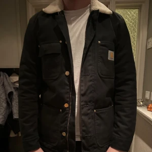Svart Carhartt jacka med ull - Svart jacka från Carhartt med ull vid kragen, guldfärgade knappar och dragkedja. Jackan har flera fickor framtill, inklusive bröstficka med Carhartt-logga. Perfekt för dig som gillar en clean look. Använd fåtal gånger och säljer på grund av att den inte kommer till användning. Den har inga defekter.