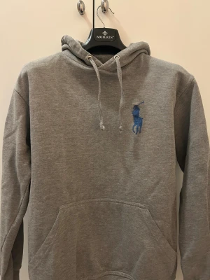 Grå hoodie från Ralph Lauren - Säljer en stilren grå hoodie från Ralph Lauren med blå logga på bröstet och stort nummer 3 på ärmen och ryggen. Tröjan är bra skick förutom att den är lite sliten på smörerna längst ut annars i bra skick!