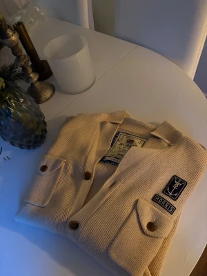 Beige Morris kofta  - Snygg beige cardigan kofta från Morris i storlek XL. Jackan är stickad med fyra fickor framtill, bruna knappar och coola patchdetaljer med ankare och Morris-logga. Perfekt för dig som gillar klassisk stil med en twist. 