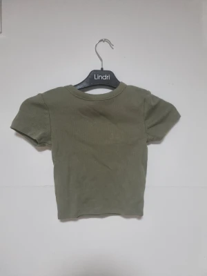 Olivgrön ribbad topp FSBN Sister XXS - Snygg olivgrön ribbad topp från FSBN Sister i storlek XXS. Modellen är croppad med rund hals och korta ärmar, perfekt för dig som gillar en enkel men trendig look. Materialet är mjukt och stretchigt, vilket gör den skön att bära hela dagen.