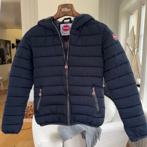 Mörkblå Colmar pufferjacka med huva - M men sitter som S.  Snygg mörkblå pufferjacka från Colmar med huva och två sidofickor med dragkedja. Jackan har ett quiltat mönster och Colmar-logga på ärmen. Perfekt för kyliga dagar och har en clean, sportig look.