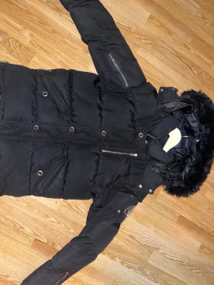 Svart dunjacka från Moose Knuckles - Säljer en svart dunjacka från Moose Knuckles med pälskantad huva och flera dragkedjor. Jackan har broderad logga på insidan och metallmärke på ärmen. Perfekt för kalla dagar och har en klassisk, stilren look.