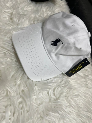 Vit keps från Polo Ralph Lauren - Snygg vit keps från Polo Ralph Lauren med klassisk broderad logga i marinblått framtill. Justerbar baktill och detaljer med Polo-tryck på insidan. Tillverkad i bomull med böjd skärm och ventilerande hål.