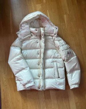 Vit Moncler pufferjacka med huva - Säljer en riktigt fet vit pufferjacka från Moncler med blank finish och stor huva. Jackan har dragkedja och knappar framtill, samt en ikonisk Moncler-logga på ärmen. Insidan har en cool serieteckning. Perfekt för kalla vinterdagar och riktigt snygg streetstil.