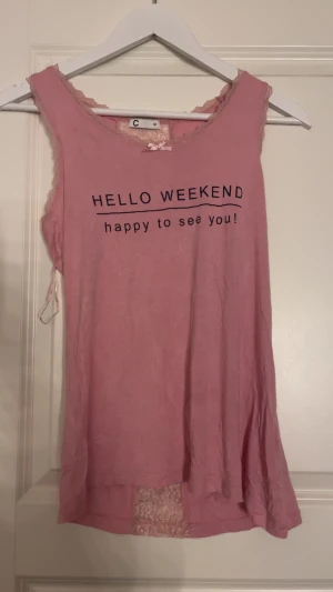 Rosa linnetopp med spets och text - Säljer en söt rosa linnetopp från C i storlek M. Toppen har spetsdetaljer längs halsringningen och ryggen, samt en liten rosett framtill. På framsidan står det 'HELLO WEEKEND happy to see you!'. Perfekt för dig som gillar mjuka färger och fina detaljer.