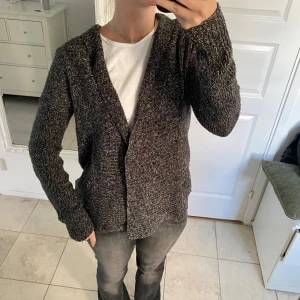 Mörkgrå stickad kofta med v-ringning - Säljer en mörkgrå stickad kofta med v-ringad öppning och långärmad passform. Koftan har en grovstickad struktur med inslag av ljusare trådar som ger en snygg melerad look. Perfekt att slänga över en t-shirt för en chill och avslappnad stil.