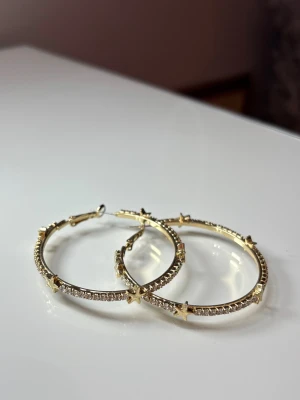 Guldiga hoops med stjärnor och stenar - Säljer ett par stora, runda örhängen i guldfärgad metall med gnistrande stenar runt hela ringen och små stjärnor som dekoration. Perfekt för dig som gillar att sticka ut med dina accessoarer och vill ha lite extra bling.