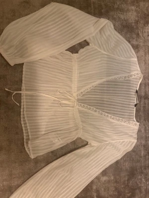 Vit randig blus med spets och knyt - Säljer en vit blus med tunna vertikala ränder, v-ringad med spetsdetaljer längs urringningen och knyt i midjan. Blusen har långa ärmar med resår vid ärmslut och är gjord i ett lätt, transparent material som ger en luftig känsla.