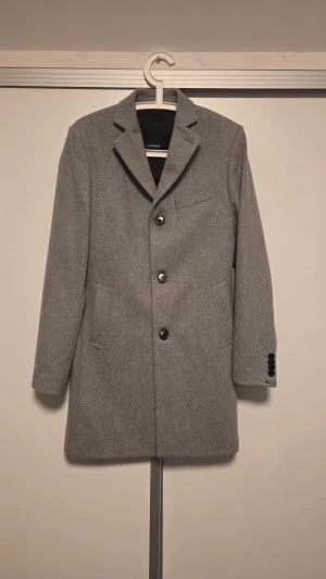 Grå ullrock från J.Lindeberg - Stilren grå rock från J.Lindeberg i ullblandning med klassisk krage, tre svarta knappar framtill och två fickor. Fodrad med svart tyg på insidan. Perfekt för dig som vill ha en clean och tidlös look.