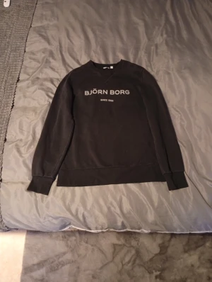 Svart sweatshirt från Björn Borg - Svart sweatshirt från Björn Borg med tryckt logga och text 'Since 1956' på bröstet. Tröjan har rund hals, långa ärmar och ribbade muddar vid ärmslut och nederkant. Perfekt för chill eller sportiga dagar.