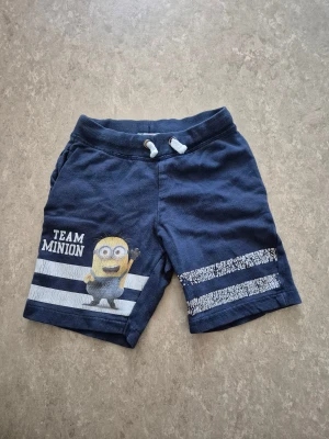 Shorts - Säljer ett par mörkblå shorts från H&M med Team Minion-tryck och coola vita ränder på benen. Shorts med snörning i midjan och mjukt bomullsmaterial, perfekt för chill dagar. Passar dig som gillar Minions och vill ha en avslappnad stil.
