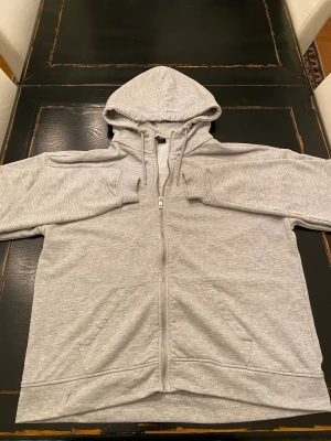 Grå zip-hoodie från SOC, storlek L - En stilren ljusgrå hoodie från SOC i storlek L. Hoodien har dragkedja hela vägen, två fickor fram och justerbar huva med snören. Tillverkad i mjukt sweatshirtmaterial.