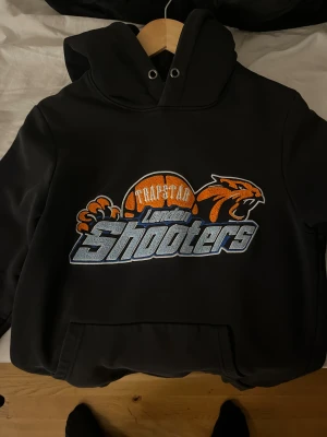 Trapstar hoodie - Svart hoodie från Trapstar med stor broderad London Shooters-logga i orange, blått och vitt på bröstet. Tröjan har huva med metallöljetter och klassisk känguruficka framtill. Perfekt streetwear-vibe med coolt baskettema.