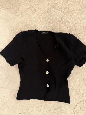 Svart blus med v-ringning Gina Tricot - Svart blus från Gina Tricot med v-ringning och korta ärmar. Toppen har snygga dekorativa knappar framtill som ger en unik touch. Figurnära passform och enkel, stilren design som passar till många olika looks.