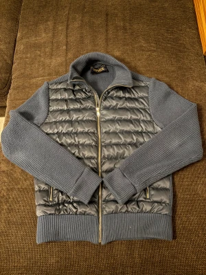 Blå pufferjacka från Vonroe - Snygg blå pufferjacka från Vonroe med stickade ärmar och rygg. Framsidan är quiltad med två dragkedjefickor och silverfärgad dragkedja. Jackan har ribbad krage, muddar och nederkant för extra komfort. Perfekt för dig som gillar stilren och modern look. Dessa är slutsålda överallt och mycket fint skick.