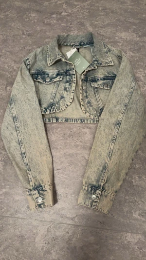 Croppad jeansjacka från H&M - Säljer en croppad jeansjacka från H&M i ljusblå tvättad denim. Jackan har klassisk krage, två bröstfickor med knappar och långa ärmar med knappslut. Perfekt för att ge din outfit en cool vibe.