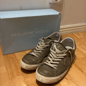 Olivgröna Philippe Model sneakers - Snygga olivgröna sneakers från Philippe Model Paris med vit sula och vita snören. Skorna har en rund tå och är tillverkade i skinn med diskreta sömmar och logodetalj på sidan. Perfekt för dig som gillar stilrena och klassiska sneakers. Dåligt skick slitna, box ingår 
