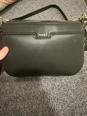 Svart axelväska från DKNY i skinn - Säljer en stilren svart axelväska från DKNY med guldfärgade detaljer och dragkedja upptill. Väskan är tillverkad i slätt skinn och har en enkel, klassisk design med justerbar axelrem. Perfekt för dig som gillar minimalistisk och snygg look. Det enda anmärkningsbara är den lilla fläcken som finns till höger om väskan 