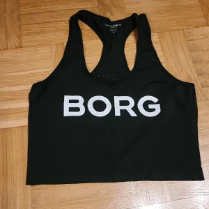 Svart sporttopp från Björn Borg - Svart sportig linnetopp från Björn Borg med stort vitt BORG-tryck framtill. Modellen har brottarrygg och små ventilationshål för extra andningsförmåga. Perfekt för träning och aktiva dagar. Tillverkad i stretchig polyester för skön passform.