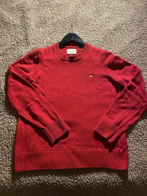 Röd stickad tröja från GANT - Säljer en röd stickad tröja från GANT med rund hals och broderad logga på bröstet. Tröjan har långa ärmar och breda ribbade muddar vid ärmslut och nederkant. Perfekt för dig som gillar klassisk stil med en modern touch.  Väldigt FIN HELT NY !🆕 