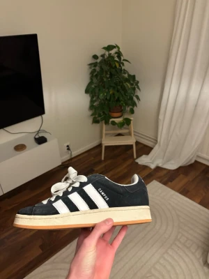 Adidas Campus sneakers svart/vit - Säljer ett par Adidas Campus sneakers i svart mocka med klassiska vita ränder och vit snörning. Skorna har en beige platt sula och rund tå. Perfekt för dig som gillar retrostil och vill ha något som funkar till streetwear.
