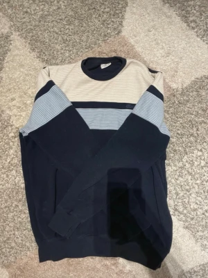 Randig Lacoste tröja slim fit XXL - Snygg långärmad tröja från Lacoste i slim fit-modell, storlek XXL. Tröjan har marinblå botten med beige och ljusblå ränder upptill och på ärmarna. Klassisk rund hals och diskret broderad Lacoste-logga på bröstet. Perfekt för dig som gillar stilrena detaljer.