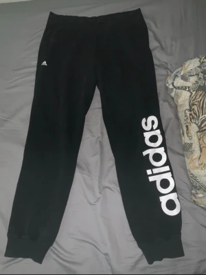 Svarta mjukisbyxor från Adidas - Svarta mjukisbyxor från Adidas med stor vit logga längs ena benet och liten logga vid fickan. Byxorna har resår och ingen snörning (lite slitna efter en tvätt som inte syns utifrån, går att sy innifrån därför är den så billig) i midjan samt ribbade muddar vid bensluten. Perfekta för chill eller träning.