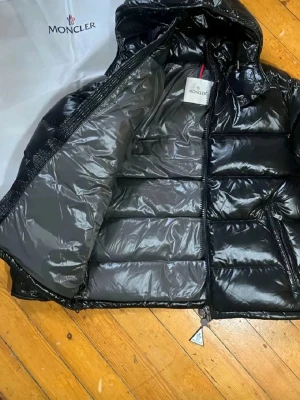 Svart Moncler pufferjacka med huva - Snygg svart pufferjacka från Moncler med glansig finish och huva. Jackan har dragkedja framtill, två fickor med dragkedja och klassisk Moncler-logga på ärmen. Perfekt för kalla dagar och ger en riktigt clean look.