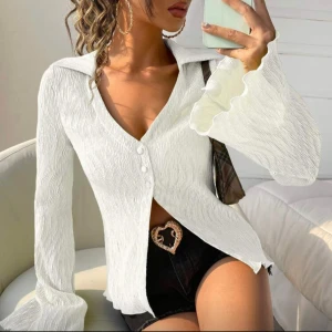 Vit blus med volangärm och v-ringning - Säljer en vit blus med v-ringning och knappar framtill. Blusen har långa volangärmar och ett lätt krinklat tyg som ger en cool struktur. Perfekt för dig som gillar trendiga detaljer och en feminin look.